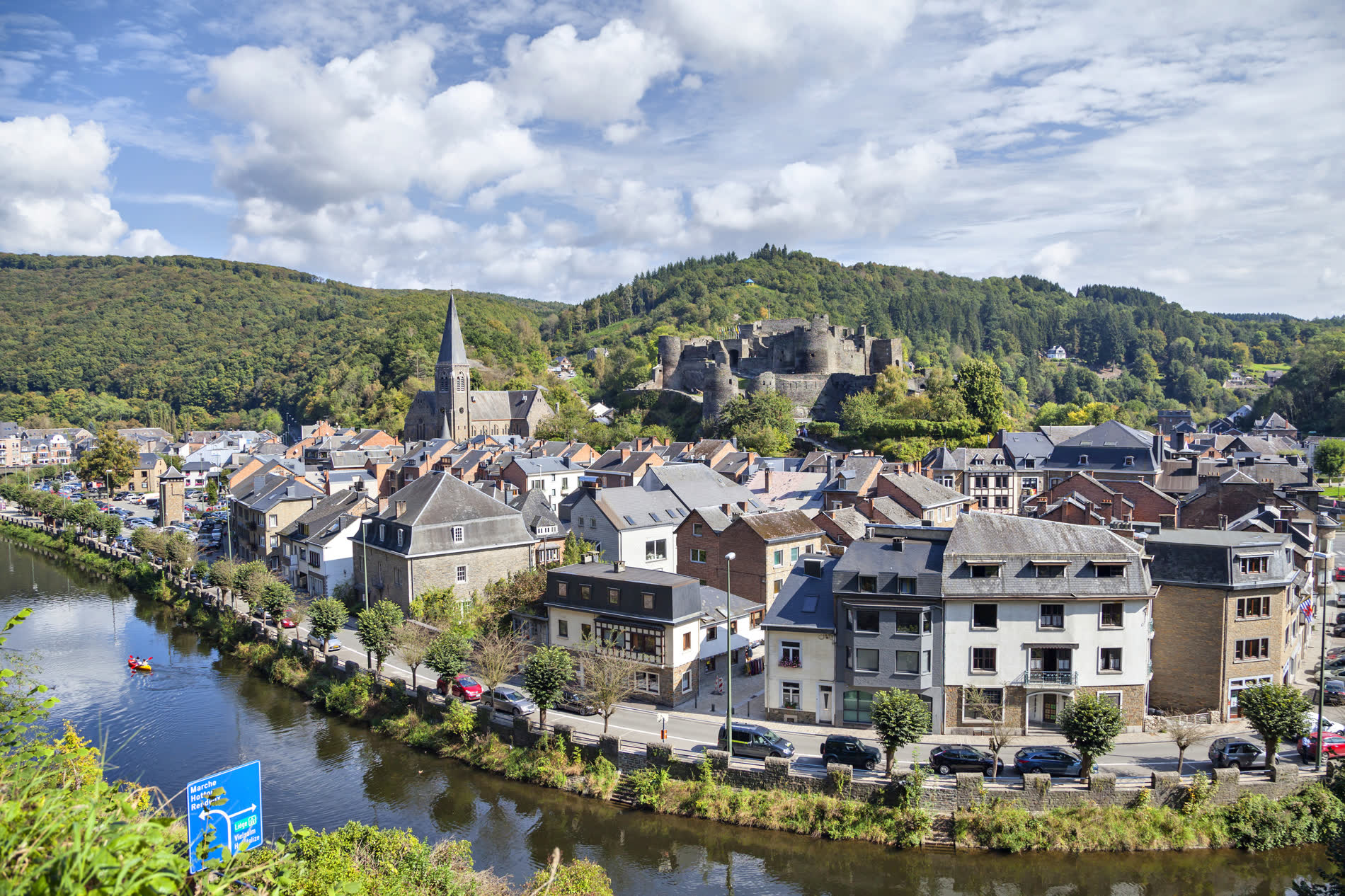Kleinstadt an einem Fluss, darüber eine Kirche und eine Burgruine am bewaldeten Hang